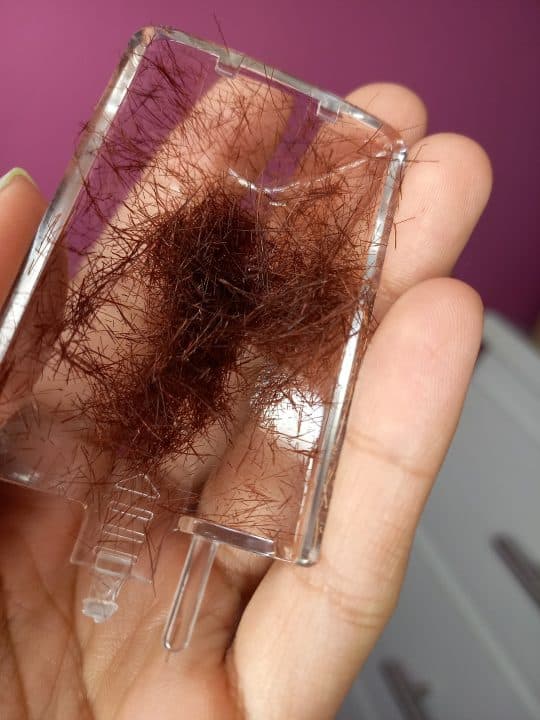 A hand holding the clear trim chamber, packed with removed brown hair ends